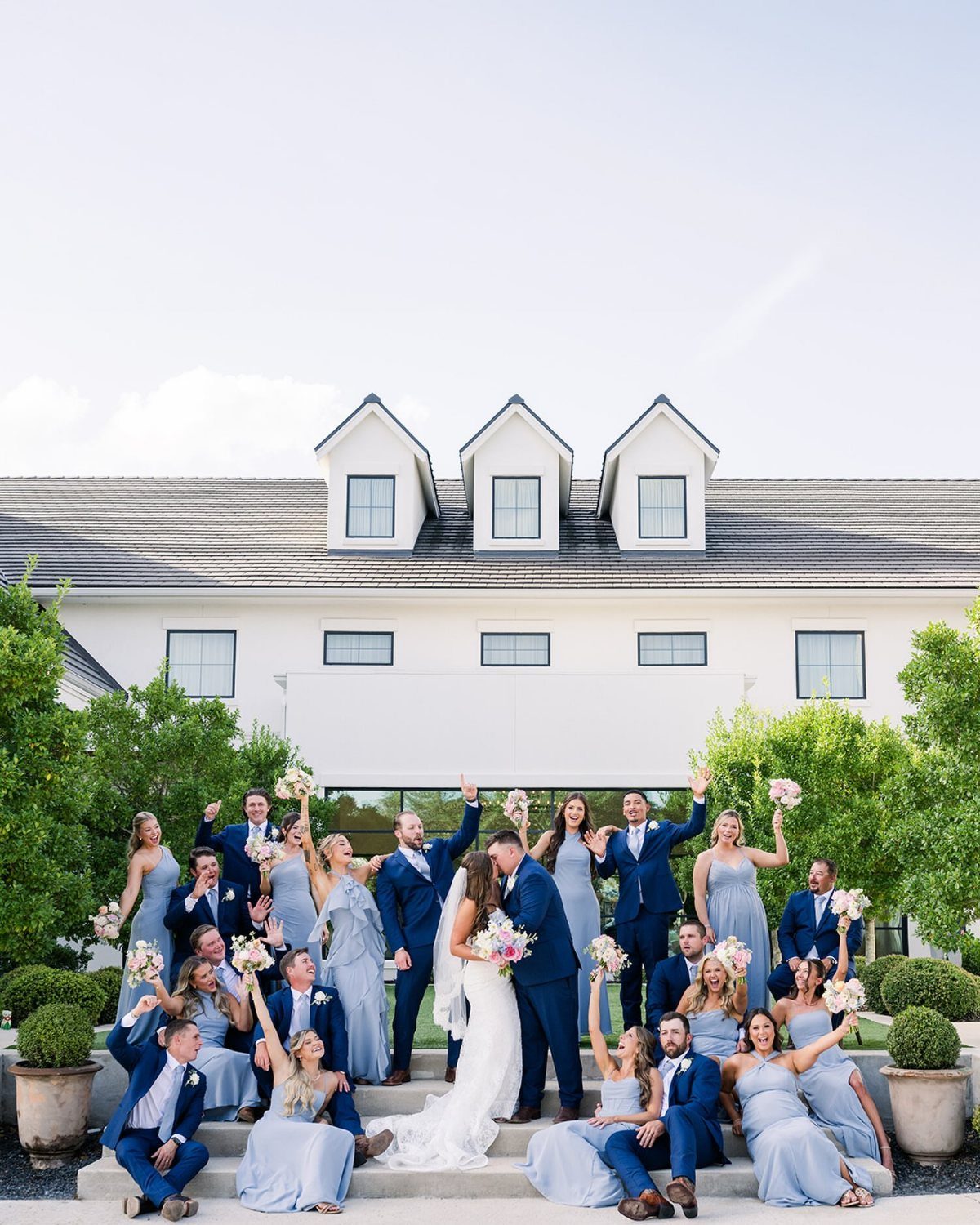 Wedding party in blue attire posing together in front of a white house.