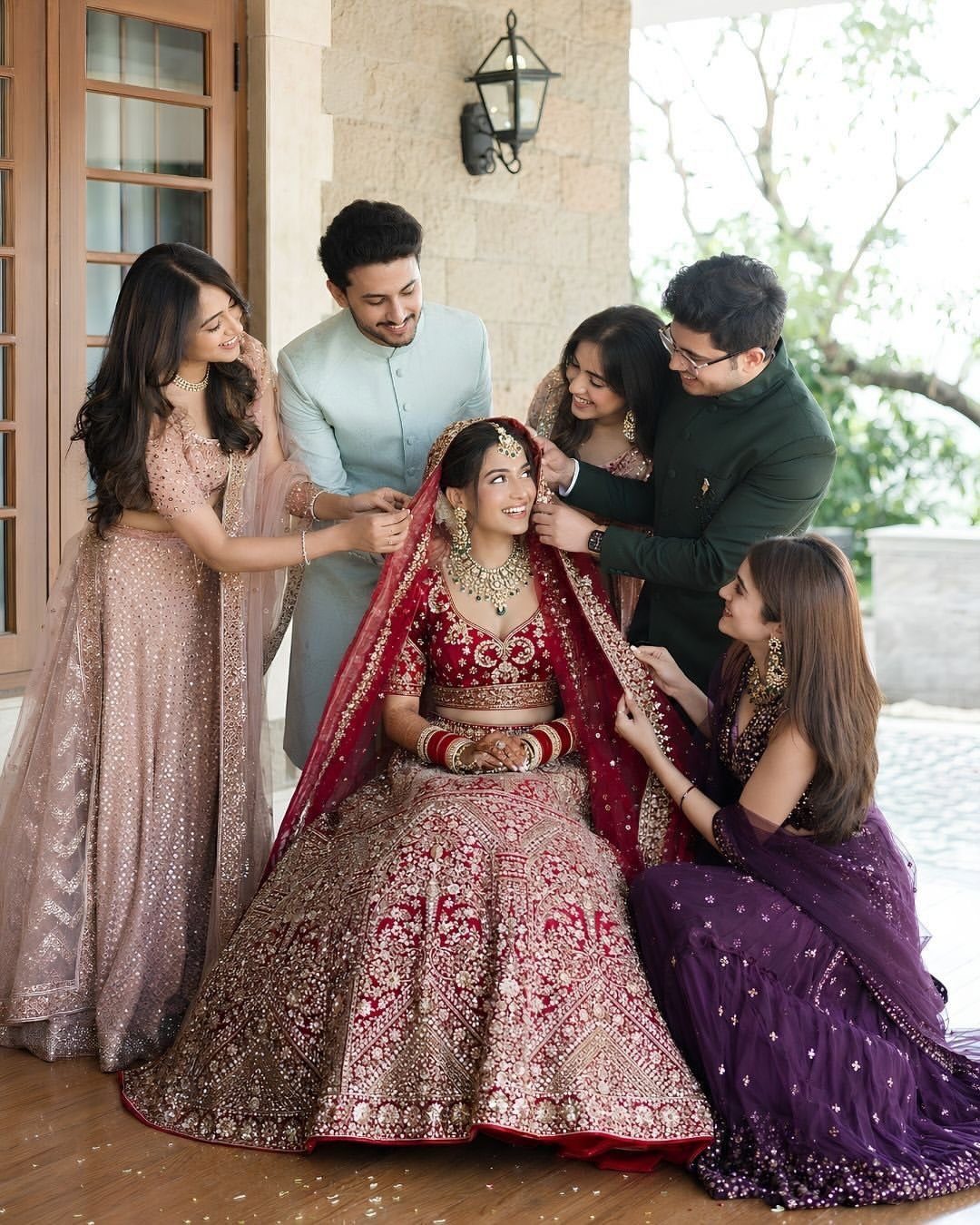 Bride in red lehenga surrounded by smiling family members in formal Indian attire.