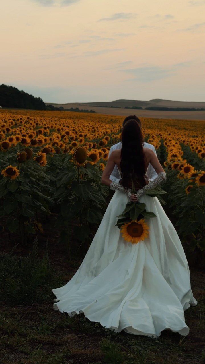 Bride and groom standing in a sunflower field during a soft golden sunset.