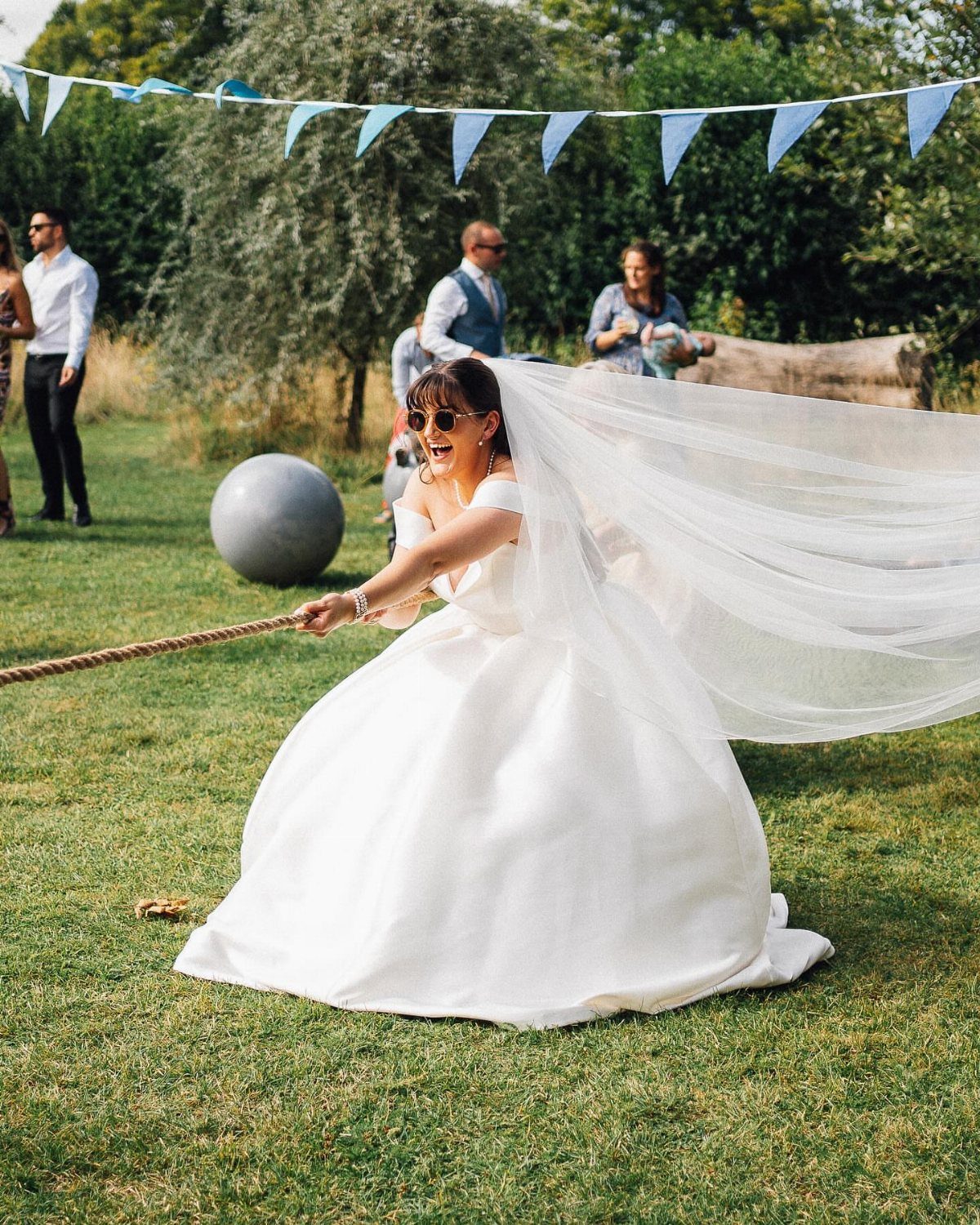 Bride in white gown pulling a rope during a tug-of-war game outdoors.