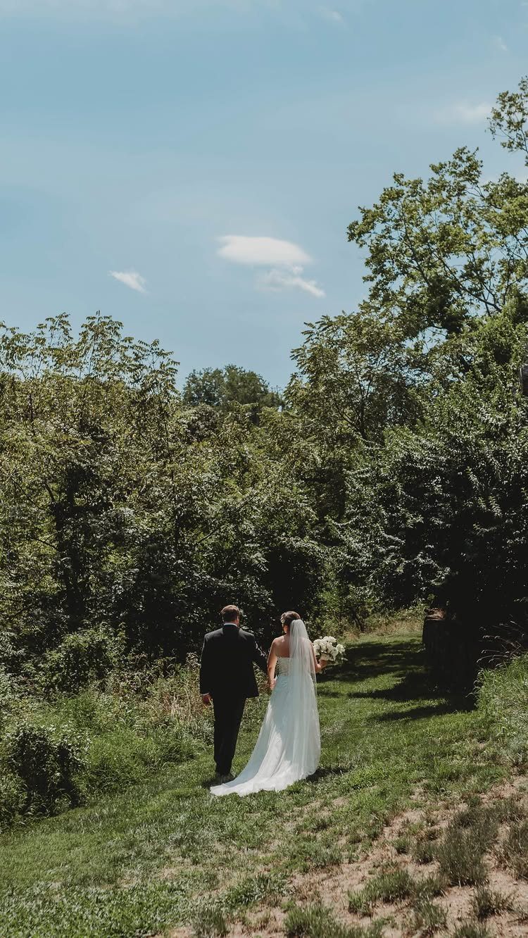 Bride and groom walking hand-in-hand away from camera through a sunny green field.