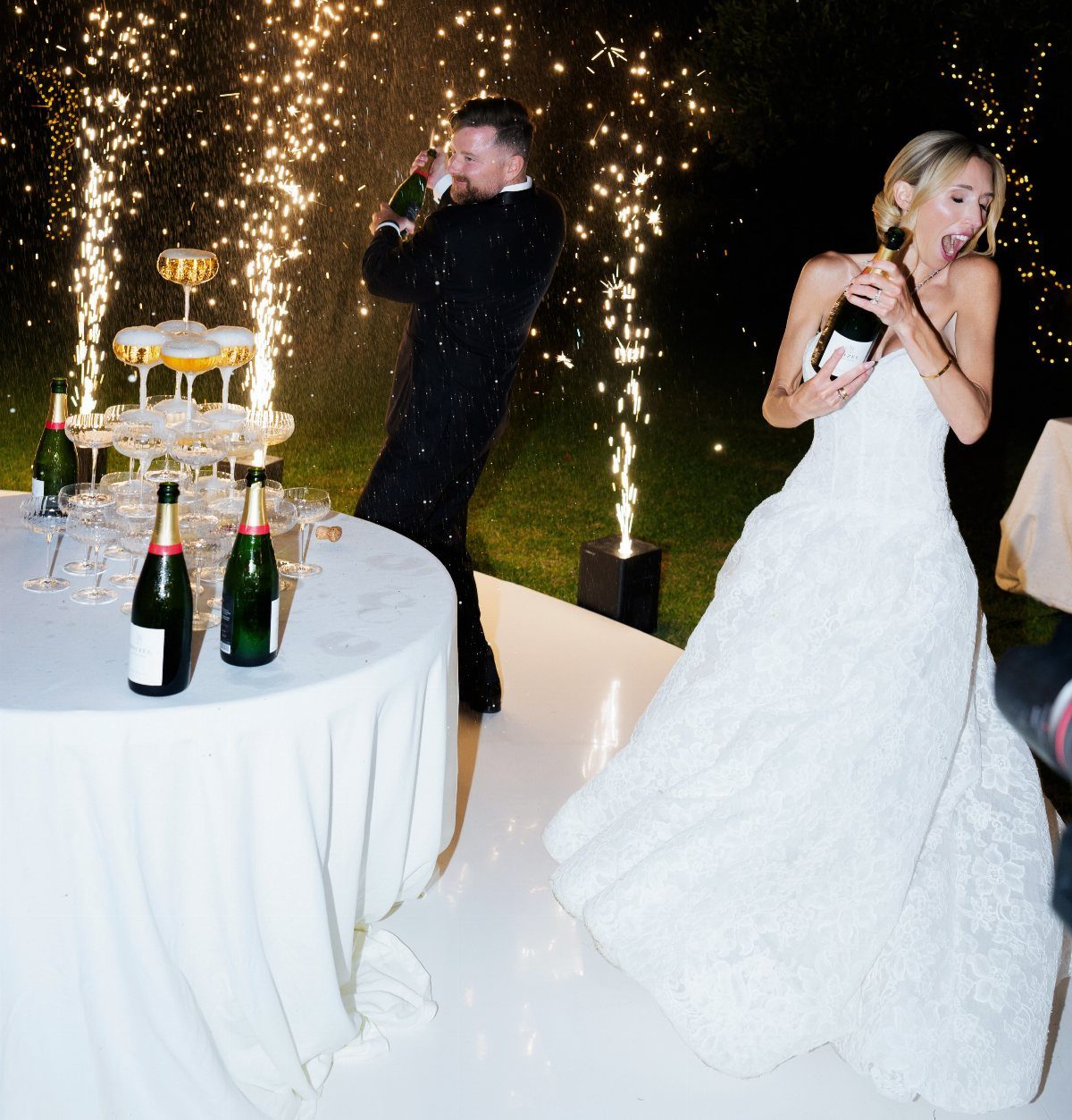 Bride and groom spraying champagne bottles next to a champagne tower with sparklers.