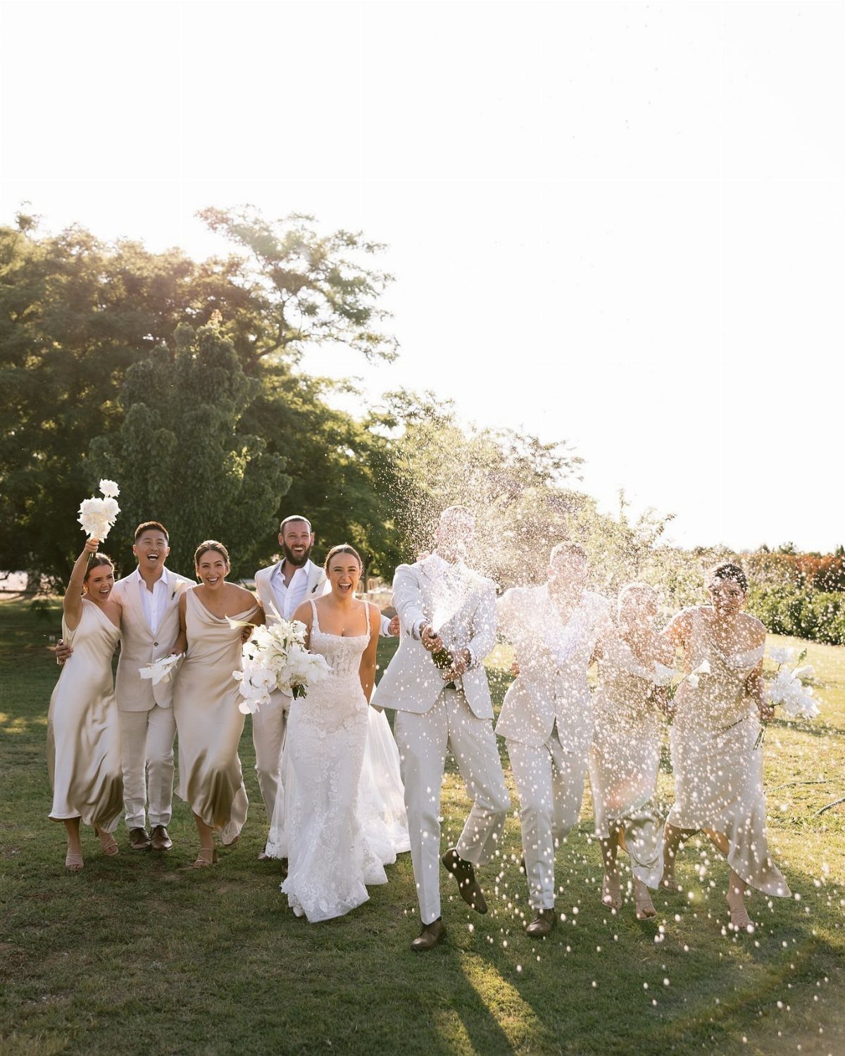 Wedding party in white walking through a field with white confetti falling around them.